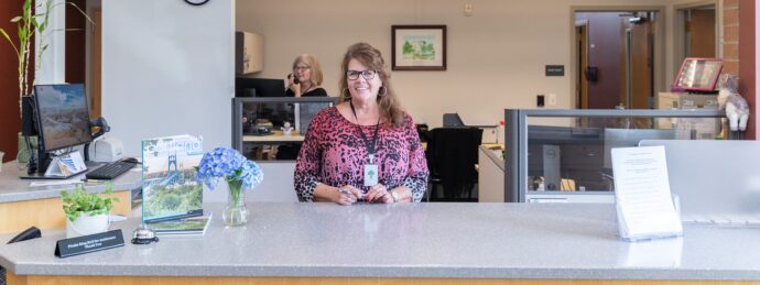 Woman in a pink and black patterned shirt smiles and stands behind the Utility Billing desk; another woman talks on the phone in the background.