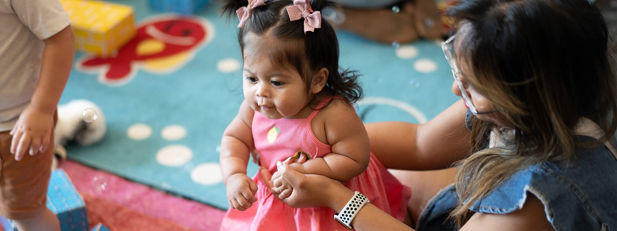 Mother holds a baby girl dressed in a pink dress upright to look at floating bubbles