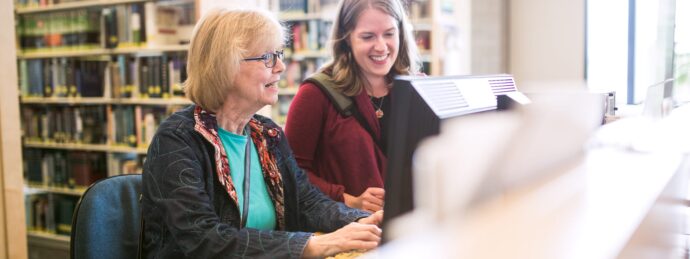 A young adult woman assists a mature woman in her 60's as she uses a public computer at the library to do some internet research.