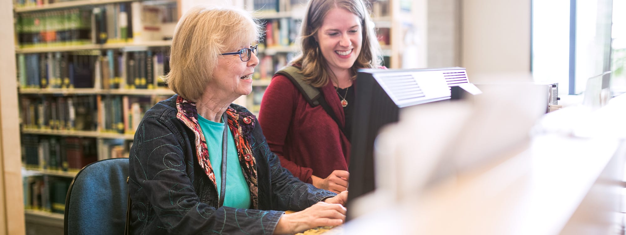 A young adult woman assists a mature woman in her 60's as she uses a public computer at the library to do some internet research.