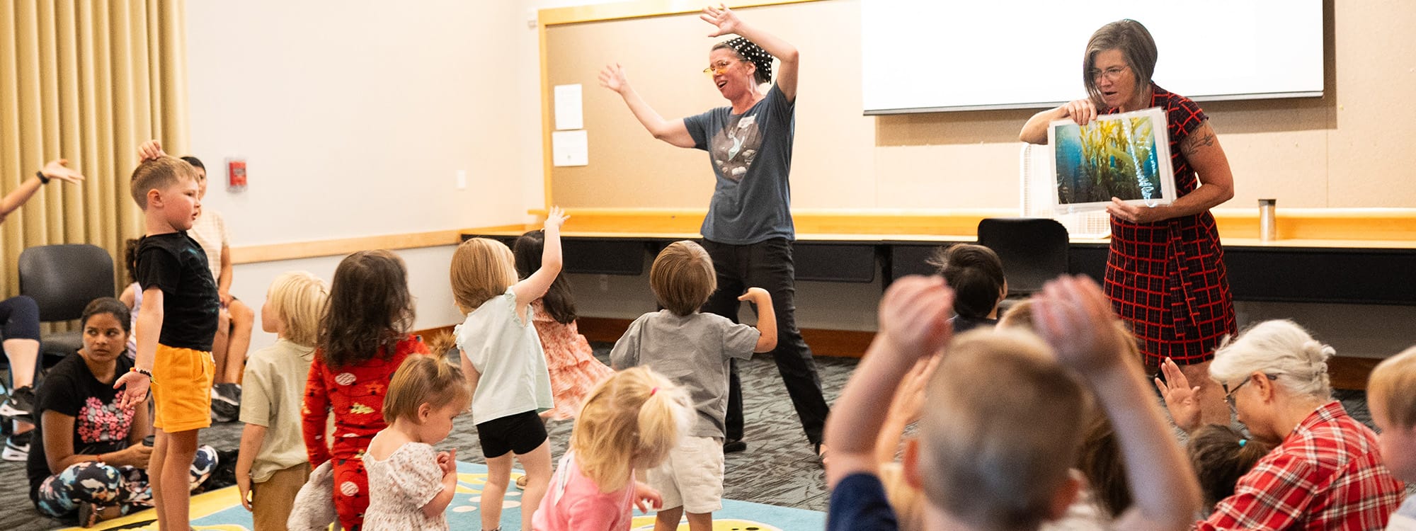 Group of young children stand and wave their arms while participating in storytime led by two women.